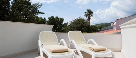 Two white lounge chairs with beige cushions on a balcony with a view of trees and a building in the background.
