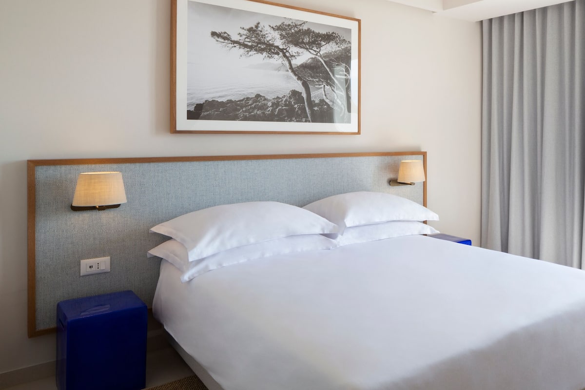 Una cama bien hecha con sábanas blancas, un cabecero con dos lámparas empotradas en la pared, un cuadro enmarcado sobre la cama y un ventanal con cortinas.