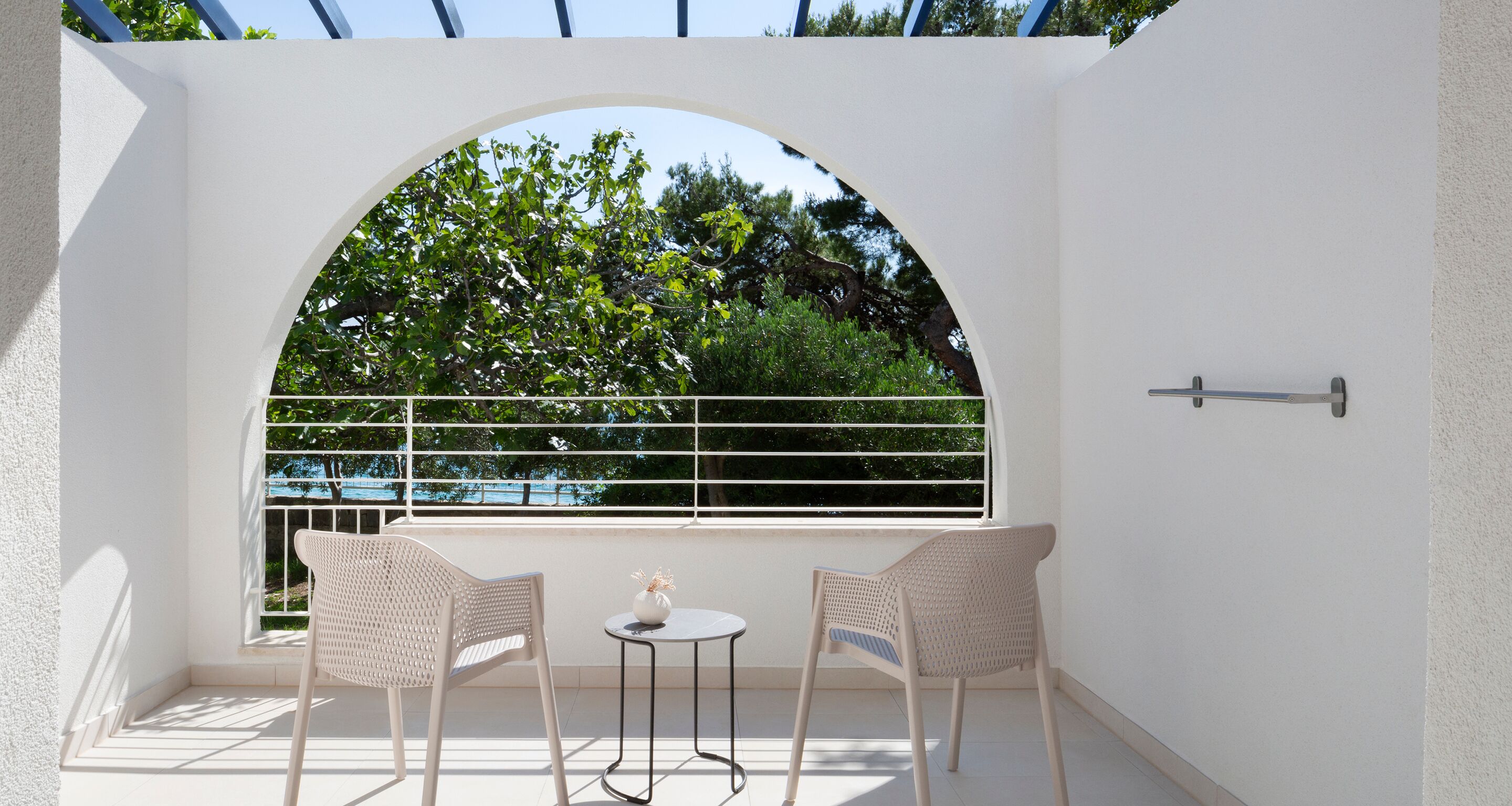 A balcony with two wicker chairs and a small table, a white railing, and a view of trees and a blue sky.