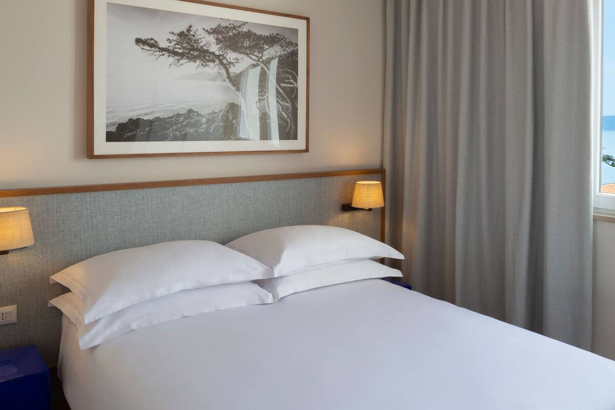 Habitación de hotel con cama, dos lámparas de noche, mesita de noche, un cuadro en la pared y vistas al mar a través de una ventana con cortinas.
