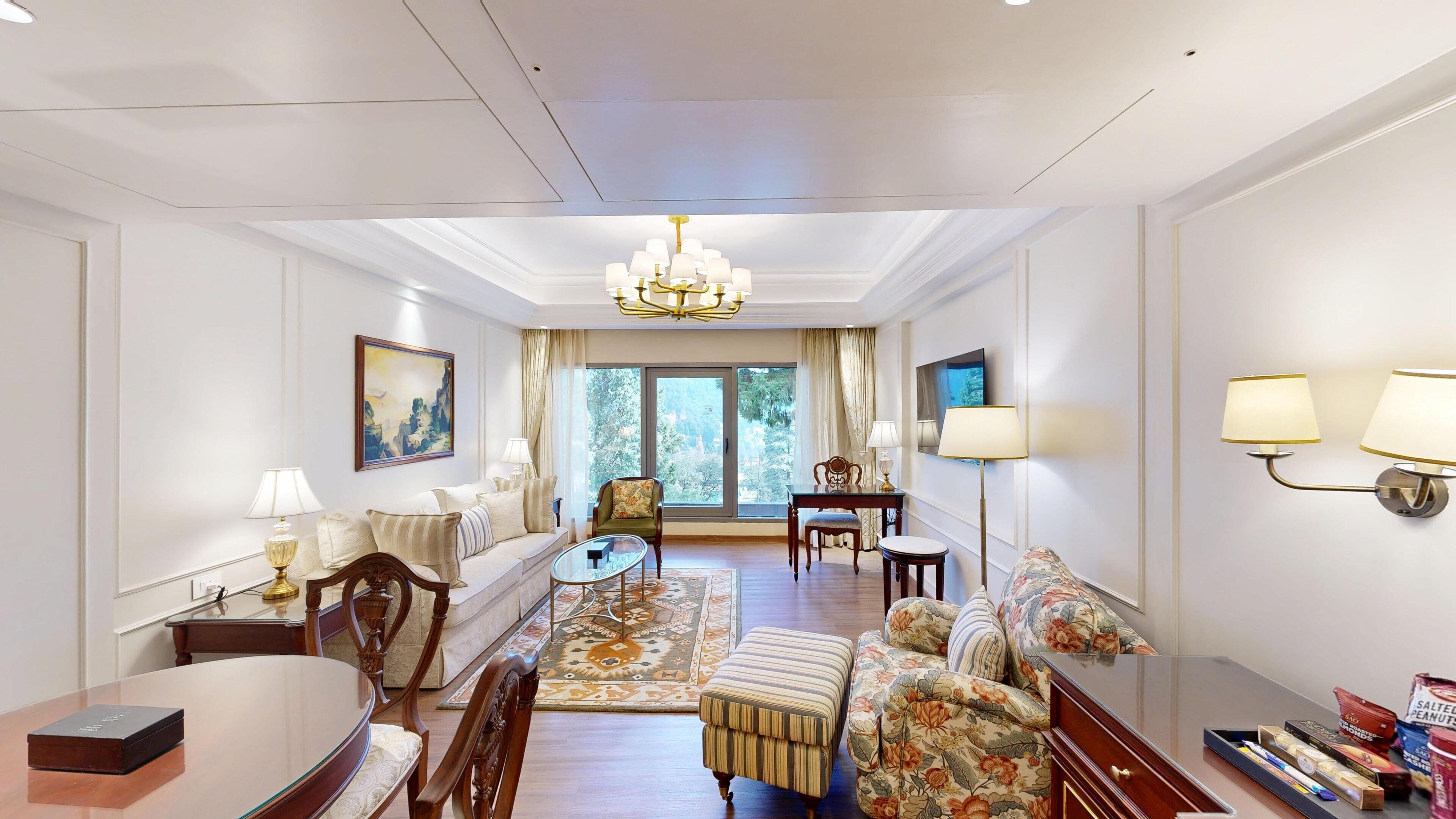 A spacious living room with a sofa, armchairs, a coffee table, and a dining table. There is a chandelier, a television, and a large window with curtains.