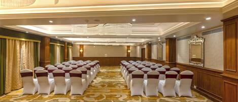 Un salón de conferencias con filas de sillas blancas adornadas con cintas color burdeos, alfombra estampada y candelabros.