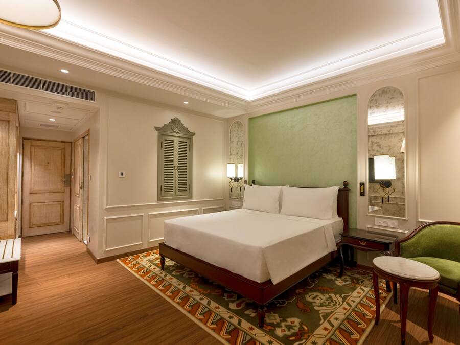 A hotel room with a large bed, a green armchair, a small table, and a wooden bench.