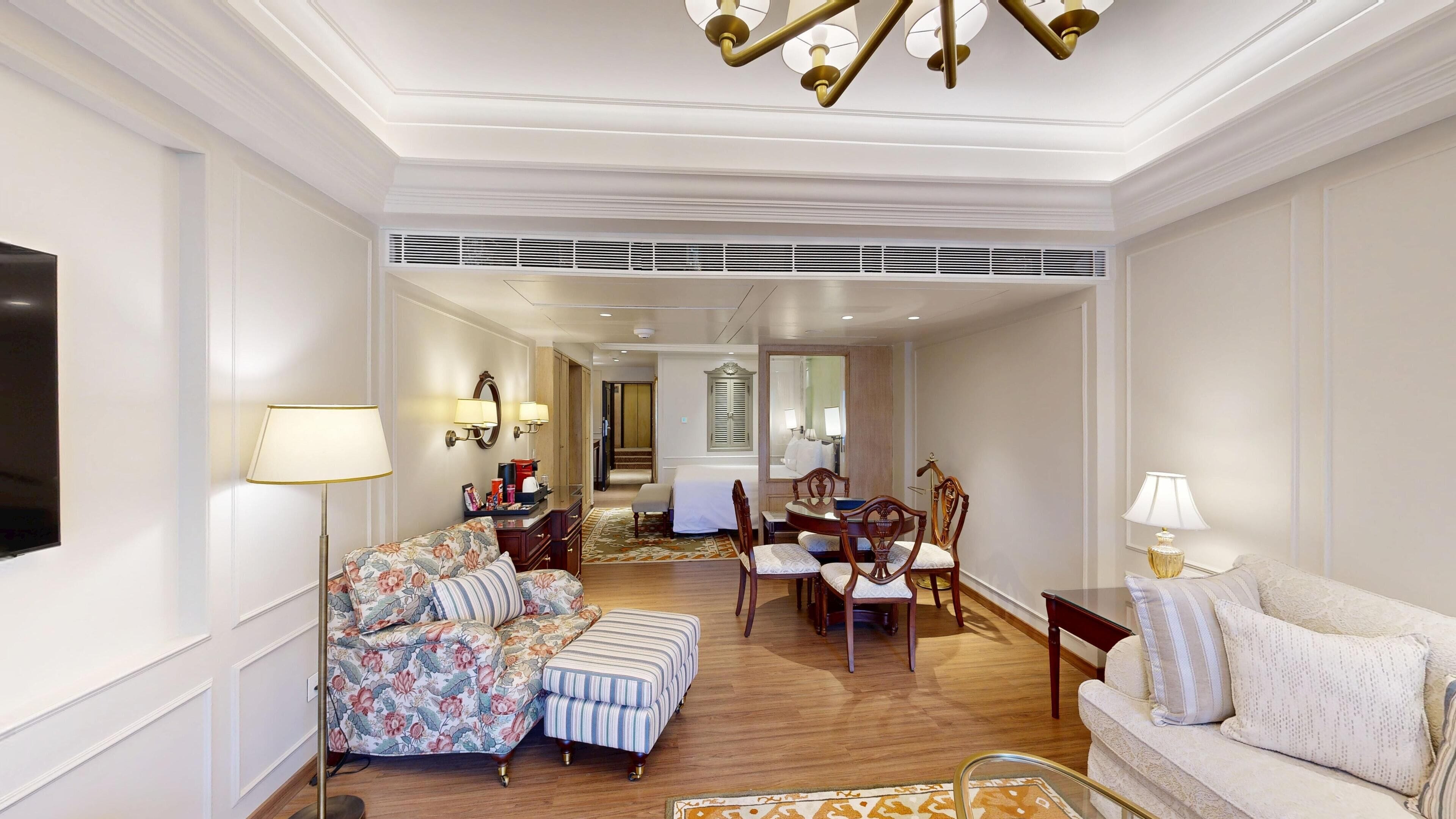 A hotel room with a sofa, ottoman, dining table, chairs, and a TV.