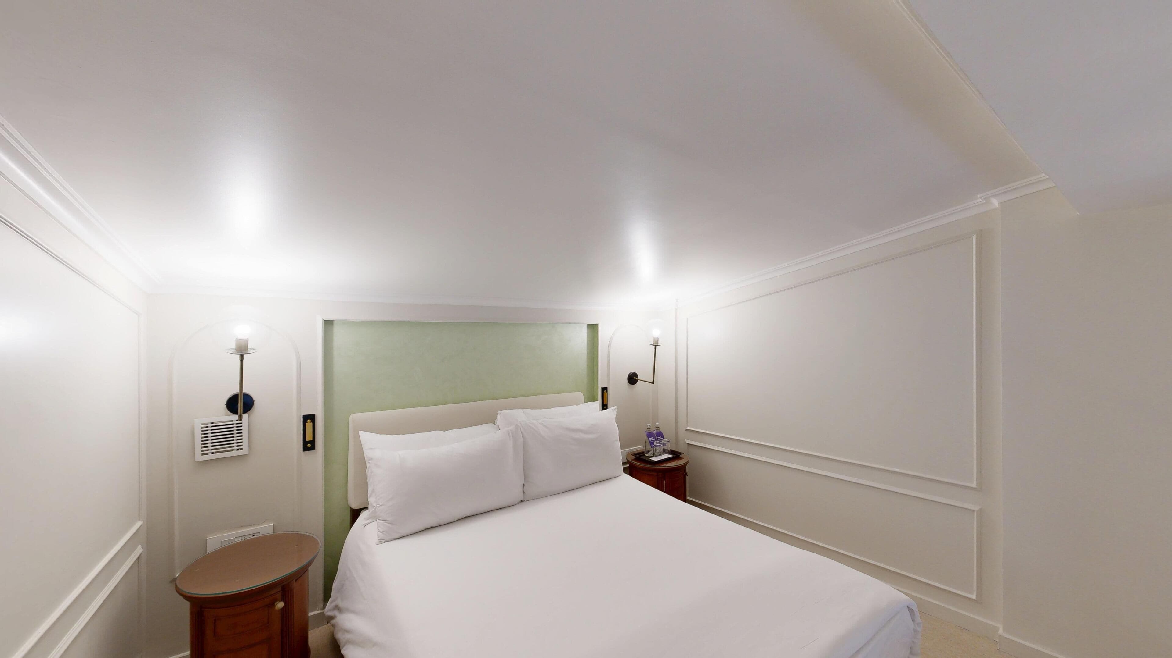 A hotel room with a large bed, a bedside table, a nightstand, a wall-mounted lamp, and a thermostat.