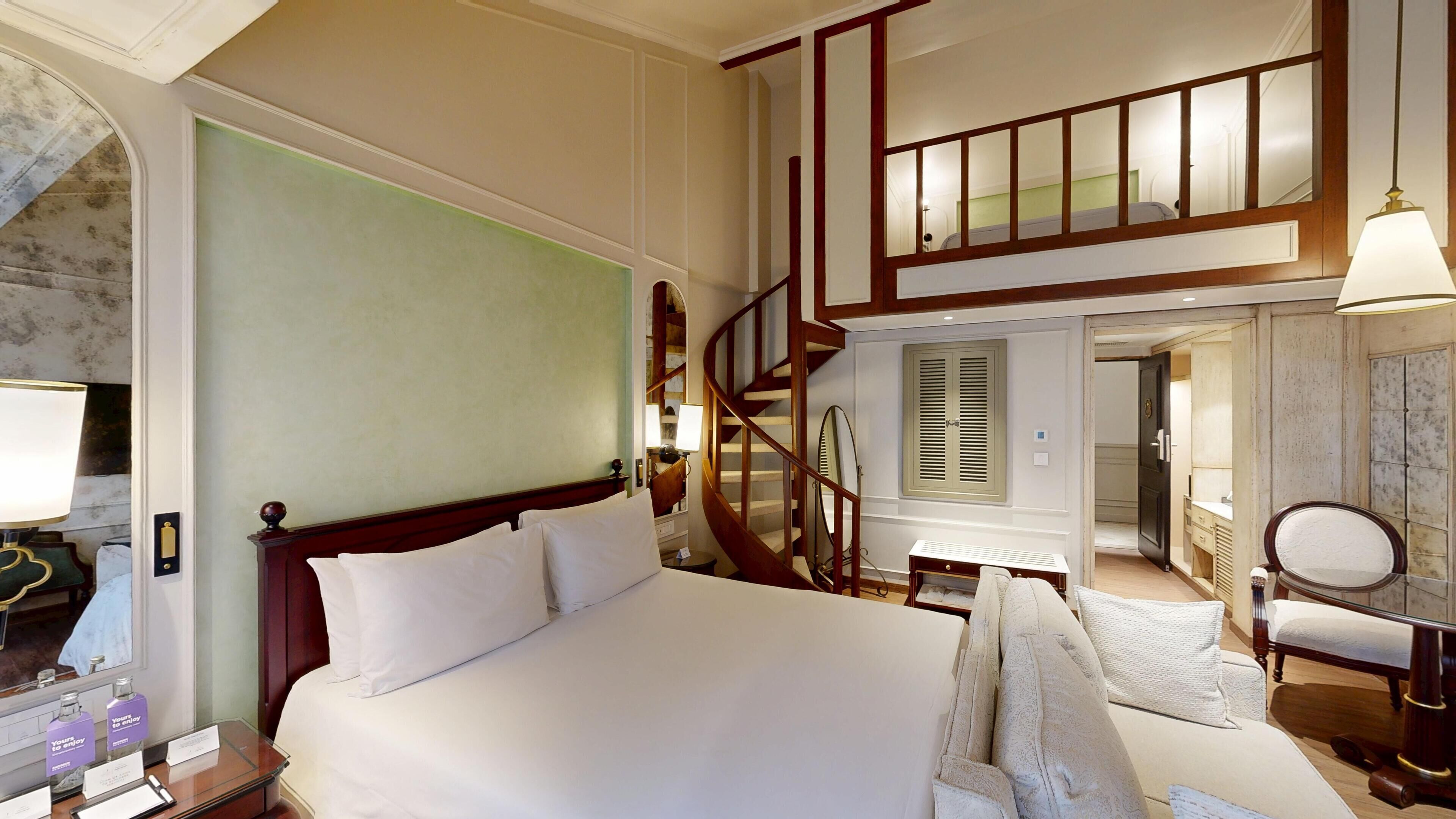 Una camera d'albergo con un letto grande, una scala in legno, un angolo salotto con una sedia e un tavolo, e un bagno parzialmente visibile attraverso una porta aperta.