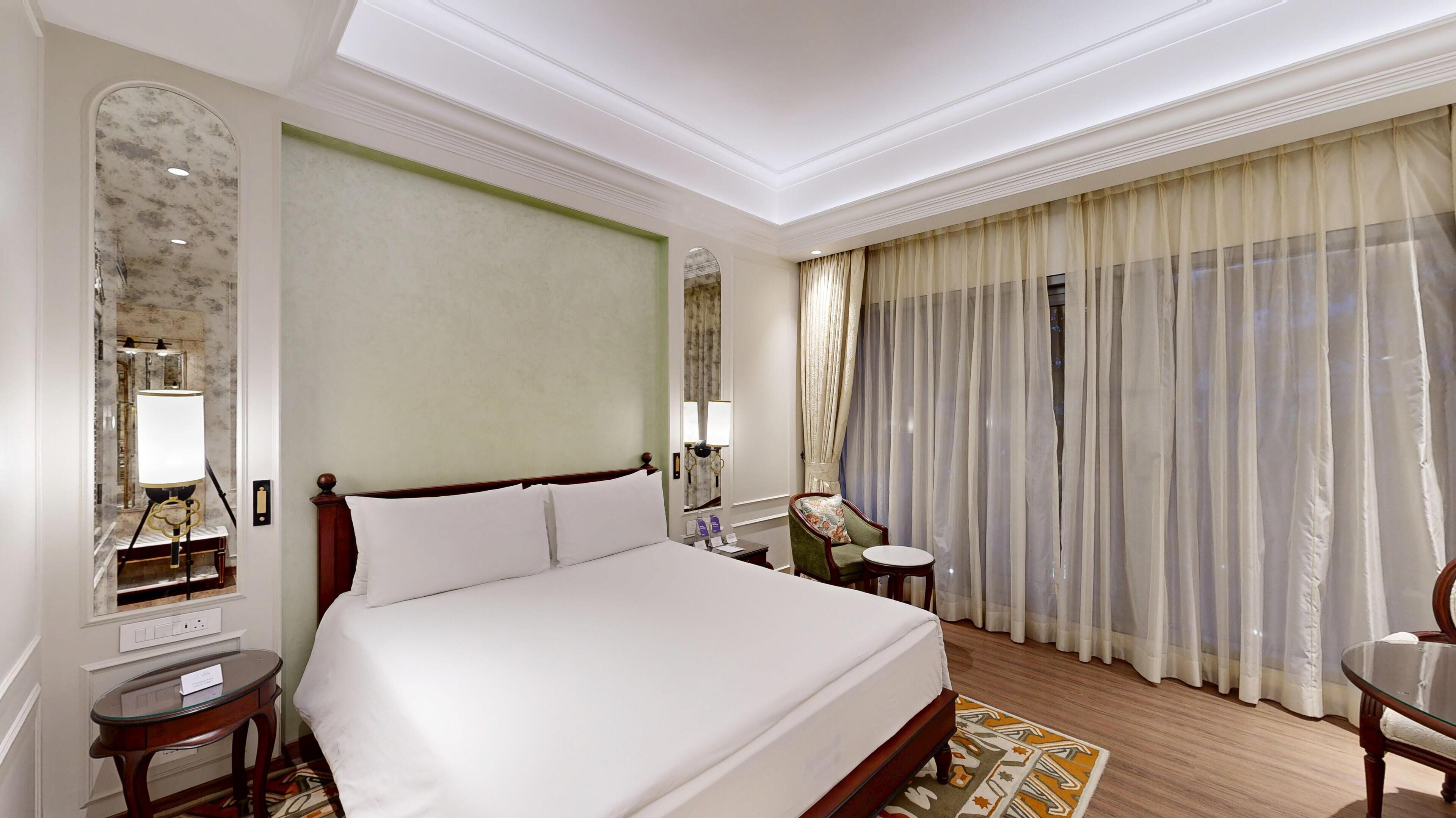 Une chambre d’hôtel avec un grand lit, un bureau, une chaise, un miroir et une fenêtre avec des rideaux.