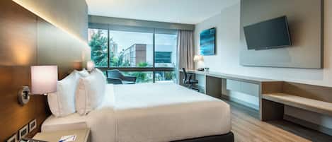 A modern hotel room with a large bed, a desk with a telephone, a flat-screen TV mounted on the wall, and a view of the outdoors through large windows.