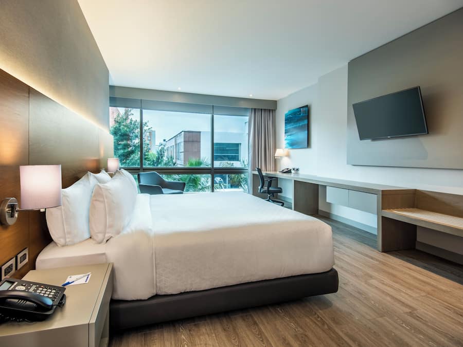 Una habitación de hotel moderna con una cama grande, un escritorio con teléfono, un televisor de pantalla plana montado en la pared y vistas al exterior a través de amplias ventanas.
