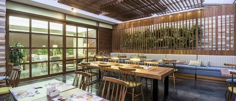 A modern dining area with wooden tables and chairs, large windows, and a wooden ceiling structure.