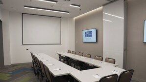 Une salle de conférence équipée d’une longue table, de chaises, d’un écran de projection et d’un écran numérique fixé au mur.