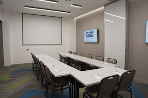 A conference room with a long table, chairs, a projector screen, and a digital display on the wall.