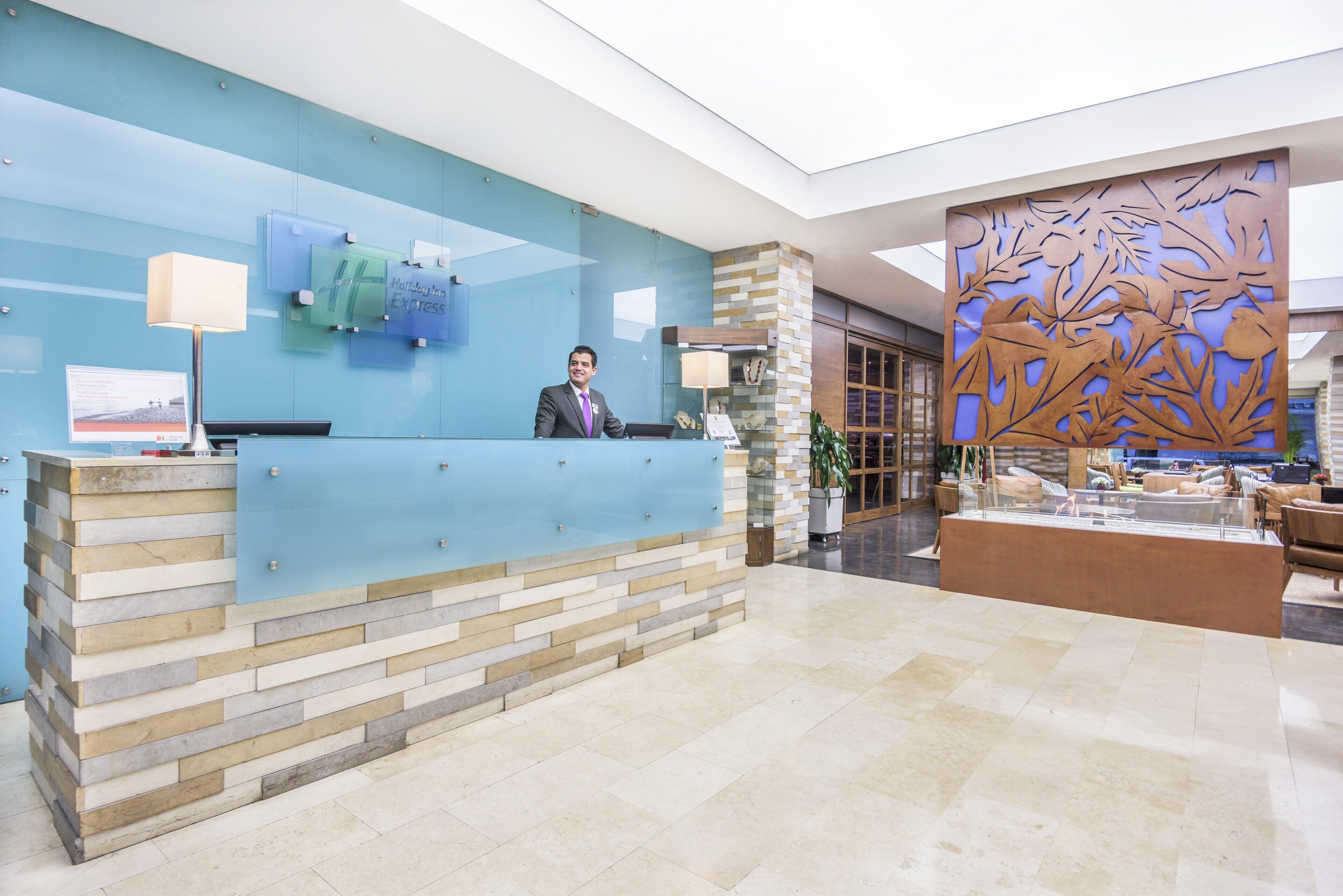 A hotel reception desk with a modern design, a receptionist, and a decorative wall with leaf patterns.