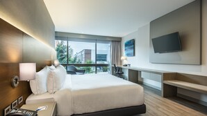 Une chambre d’hôtel moderne dotée d’un grand lit, d’une table de chevet avec un téléphone, d’un bureau avec une lampe, d’un téléviseur à écran plat fixé au mur et offrant une vue sur la ville grâce à de grandes fenêtres.