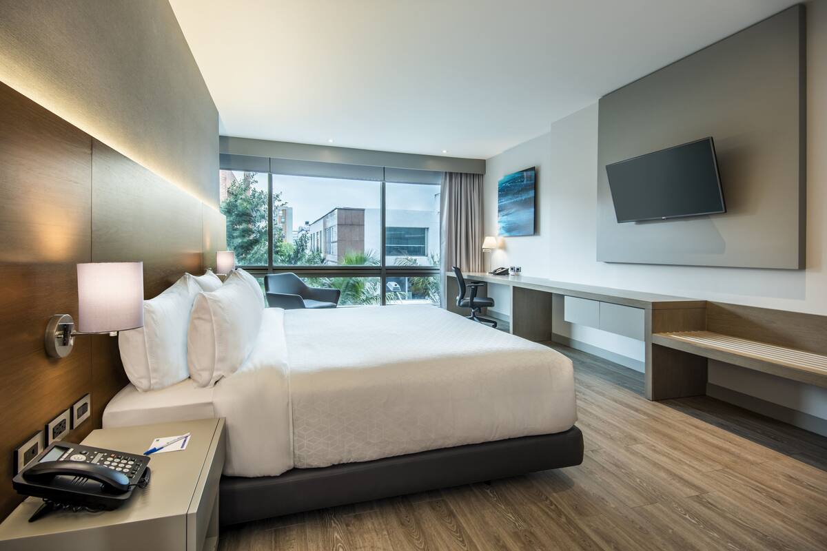 Habitación de hotel moderna con una cama grande, mesita de noche con teléfono, escritorio con lámpara, televisor de pantalla plana montado en la pared y vistas a la ciudad a través de amplios ventanales.