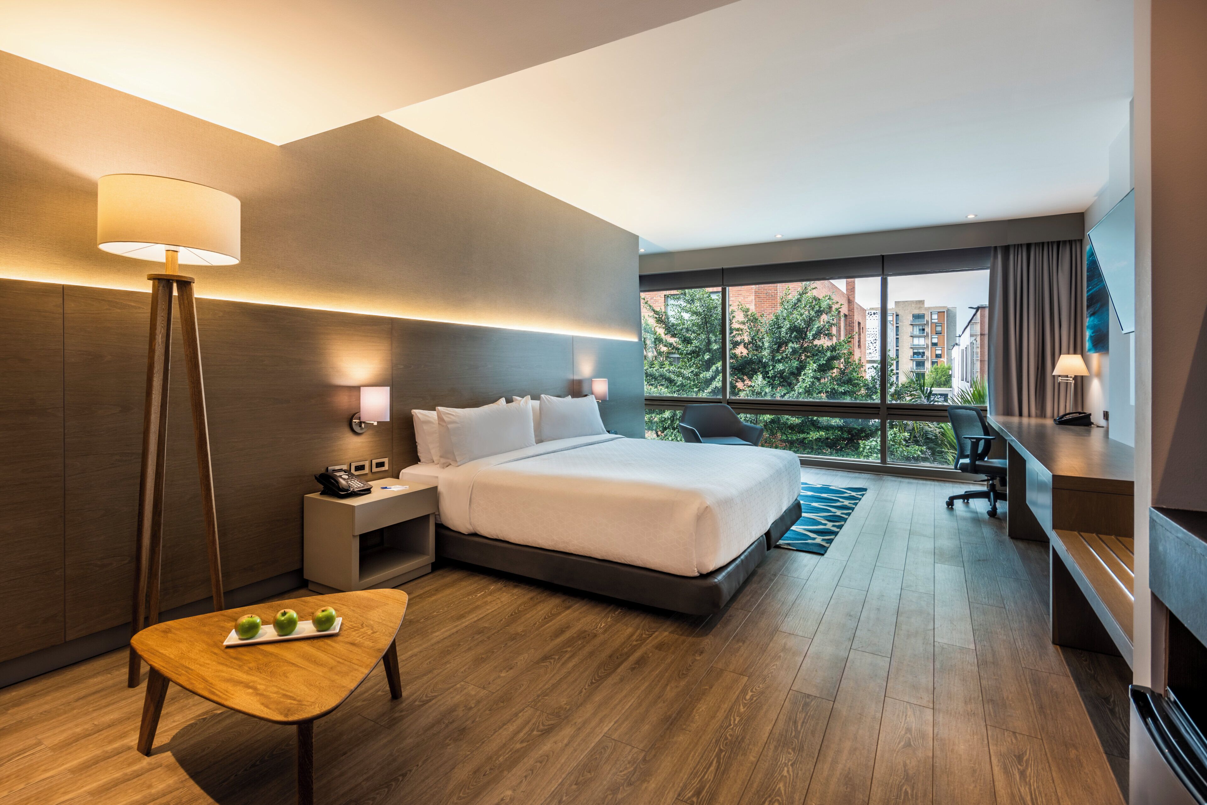 A modern hotel room with a large bed, bedside lamps, a desk, and a view of the city through large windows.