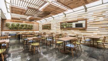 Un vaste espace intérieur aménagé en salle à manger, avec des tables et des chaises en bois, un plafond haut orné de poutres en bois et un mur décoré d’un motif géométrique.