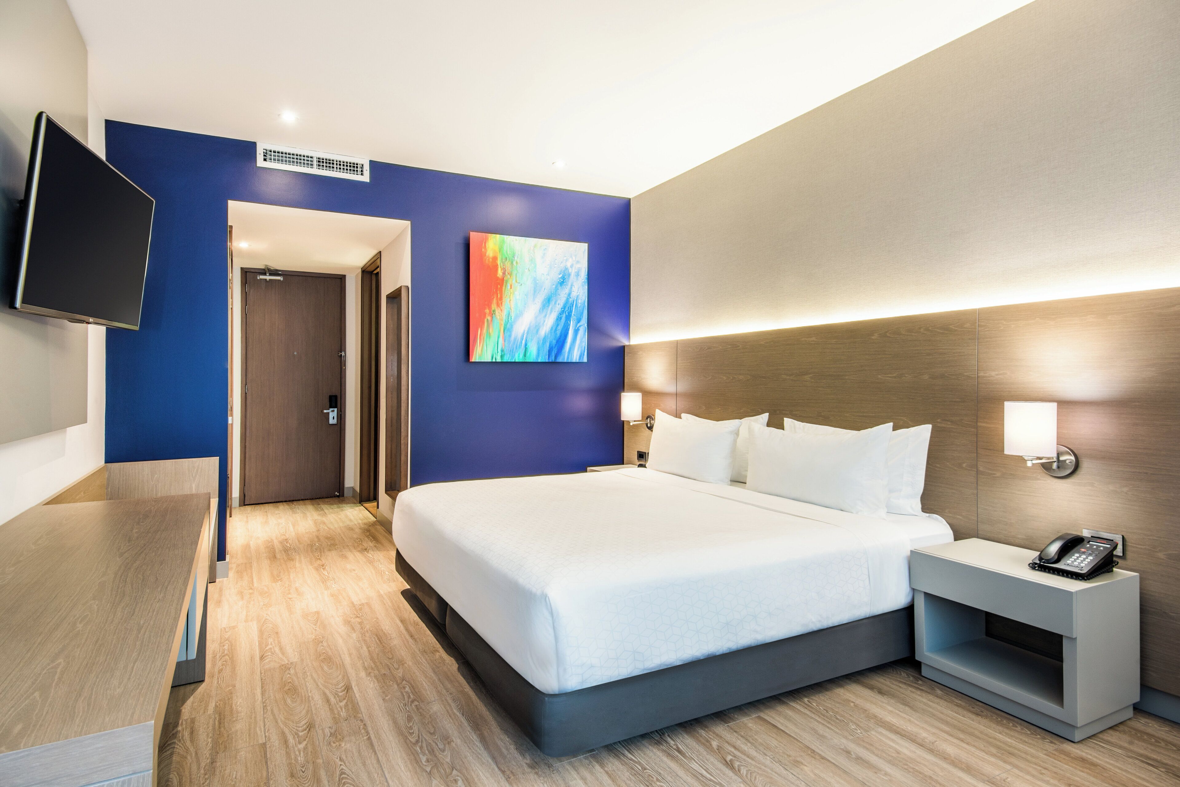 Ein Hotelzimmer mit einem Bett, einem Nachttisch, einem Telefon, einem Fernseher und einem Bild an der Wand.