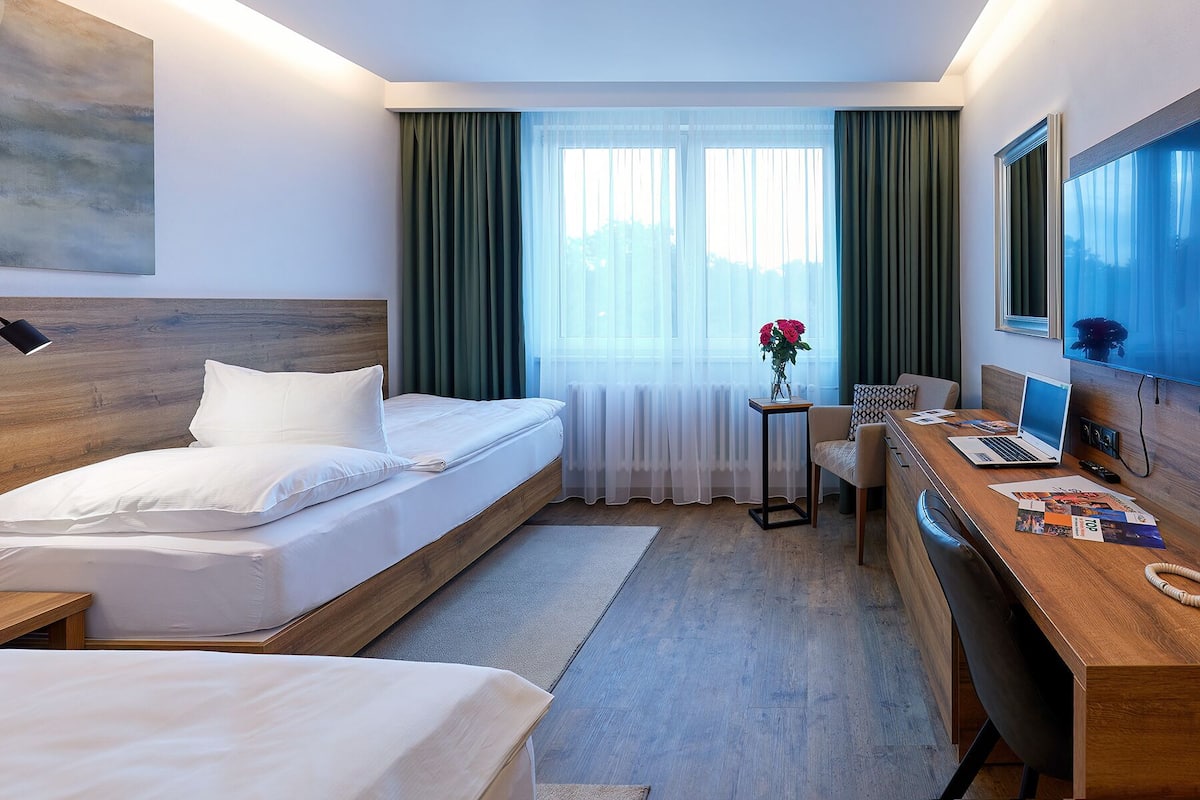 Een hotelkamer met een bed, een bureau met een laptop, een stoel en een raam met gordijnen.