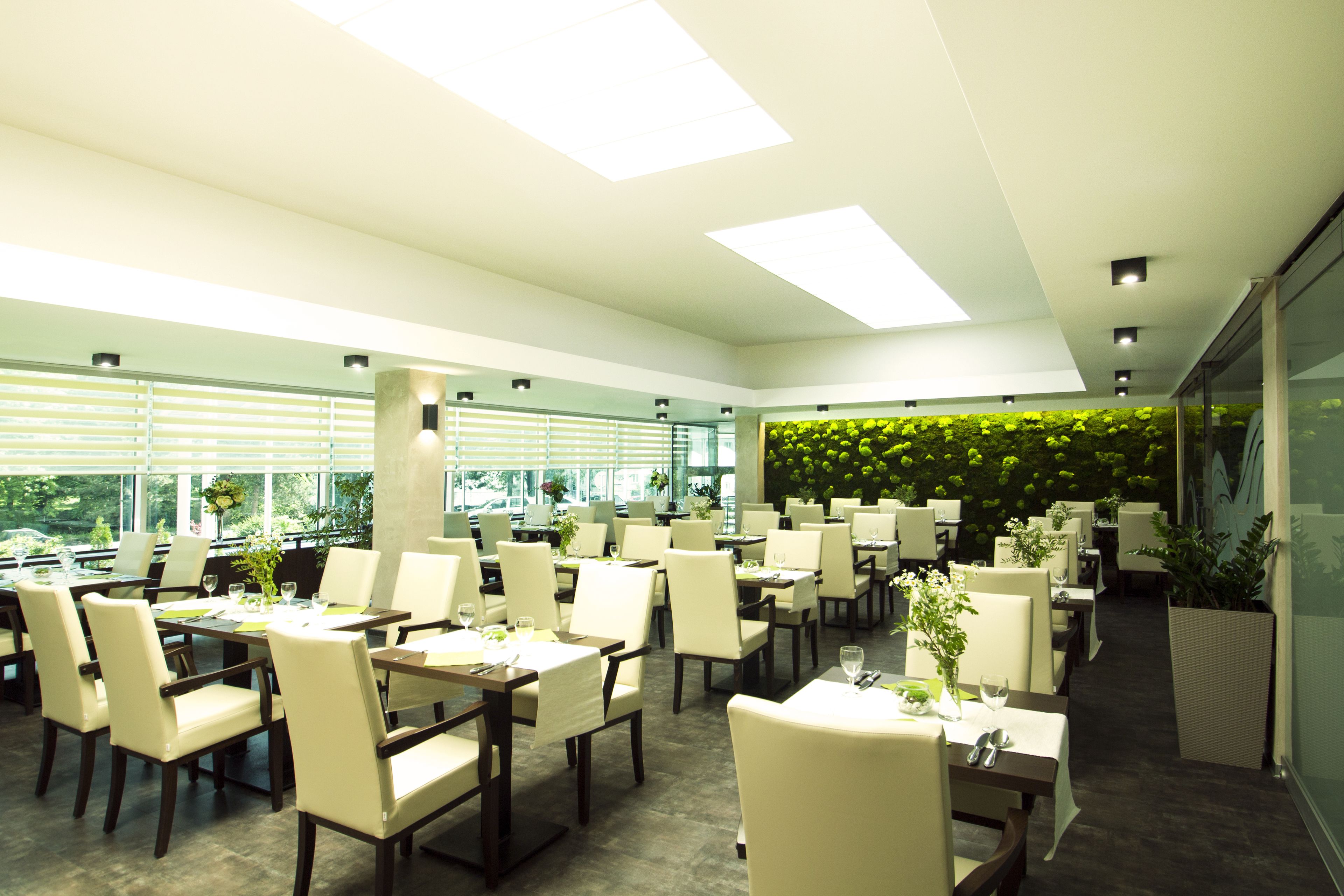 A modern restaurant with multiple tables and chairs, large windows, and a green wall with plants.