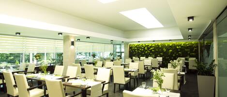 A modern restaurant with multiple tables and chairs, large windows, and a green wall with plants.