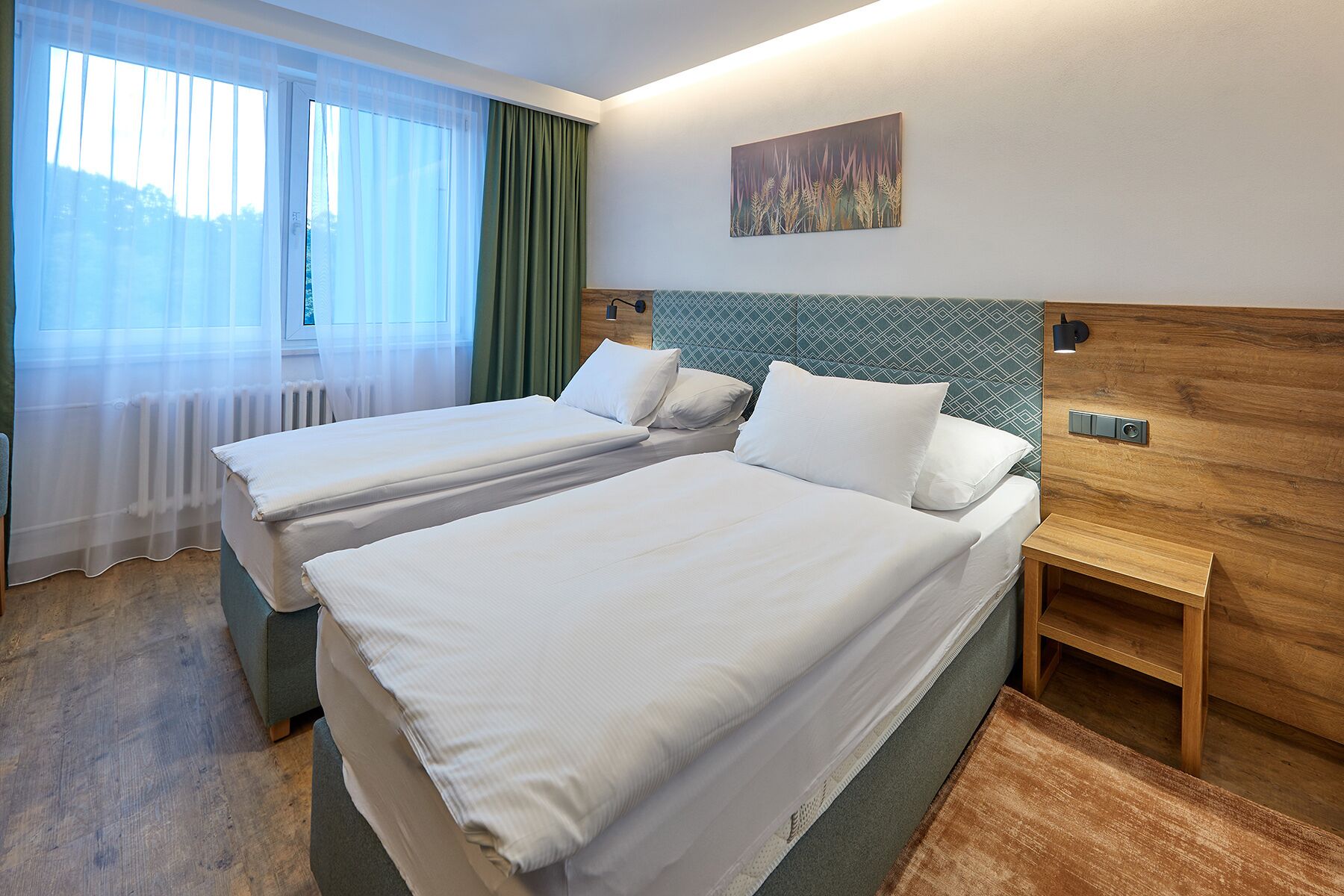 Habitación de hotel con dos camas, cabecera de madera, mesita de noche y ventana con cortinas traslúcidas.