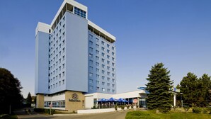 A modern hotel with a prominent corner entrance, a large parking area, and a well-maintained outdoor seating area with umbrellas. - Central Park Flora (Olomouc)