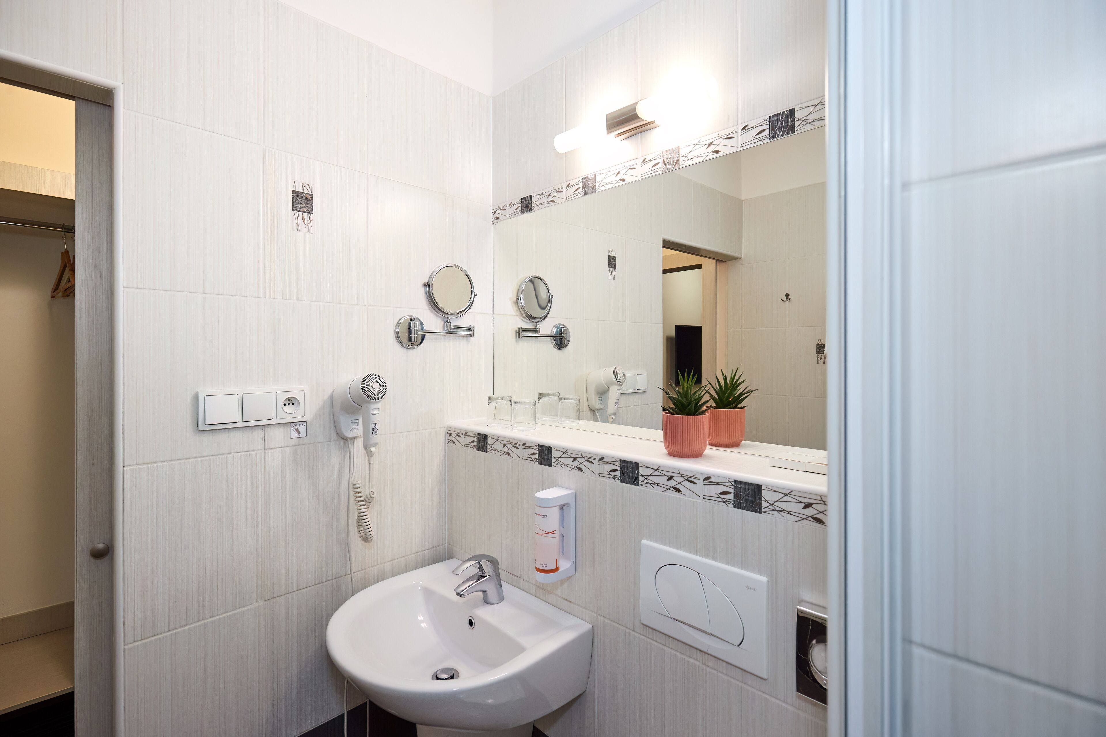 A modern bathroom with a white sink, two mirrors, a hairdryer, and a potted plant on the vanity.