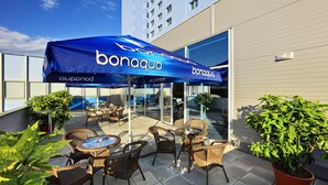 An outdoor seating area with wicker furniture and a blue umbrella branded 'bonaqua'. - Central Park Flora (Olomouc)
