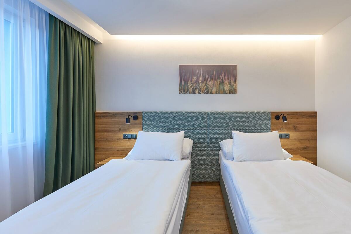 Een hotelkamer met twee bedden, een houten hoofdbord, een aan de muur hangend schilderij en een raam met groene gordijnen.