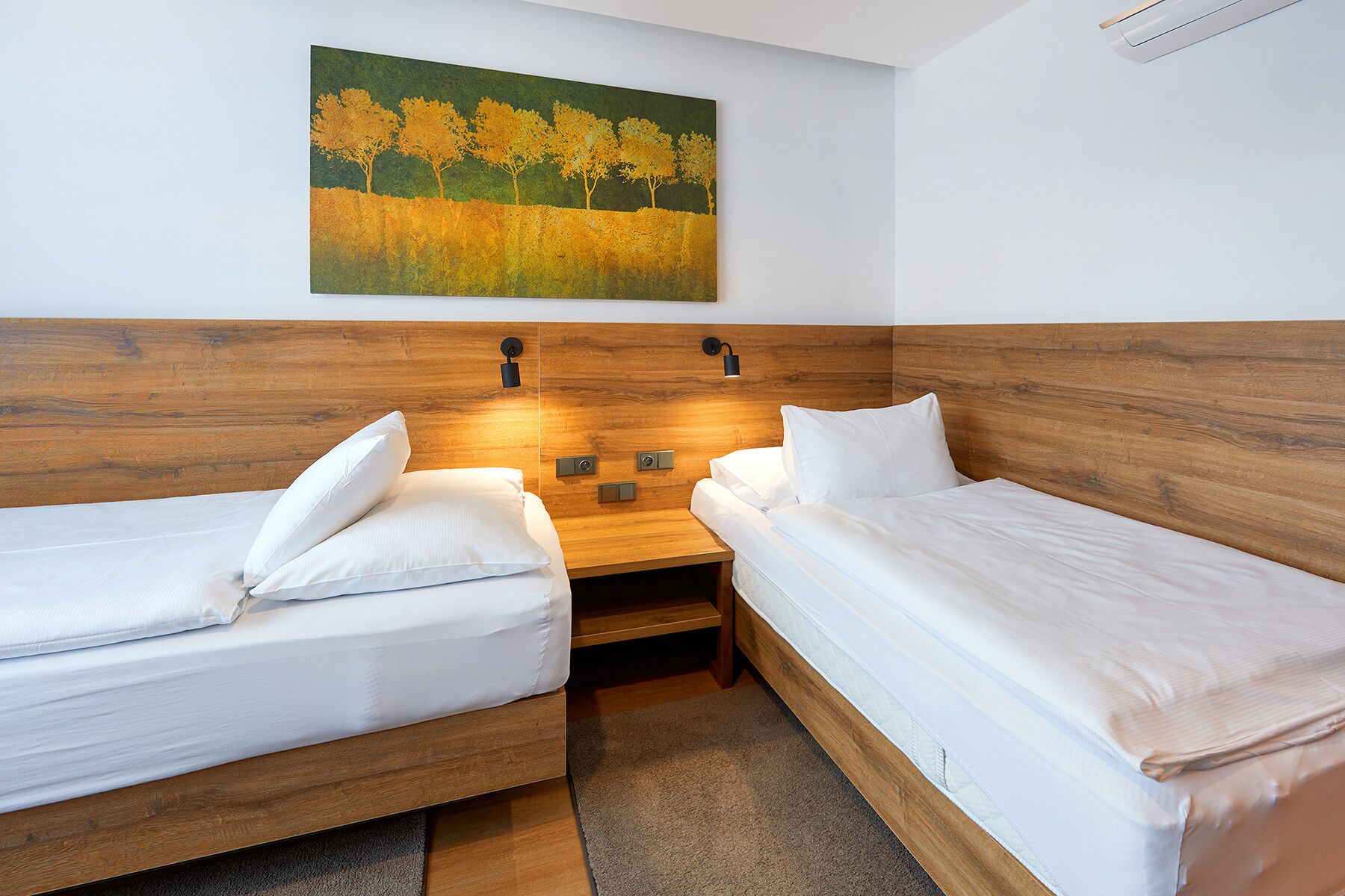 A hotel room with two single beds, a wooden headboard, a nightstand, a wall-mounted lamp, and a painting of trees.