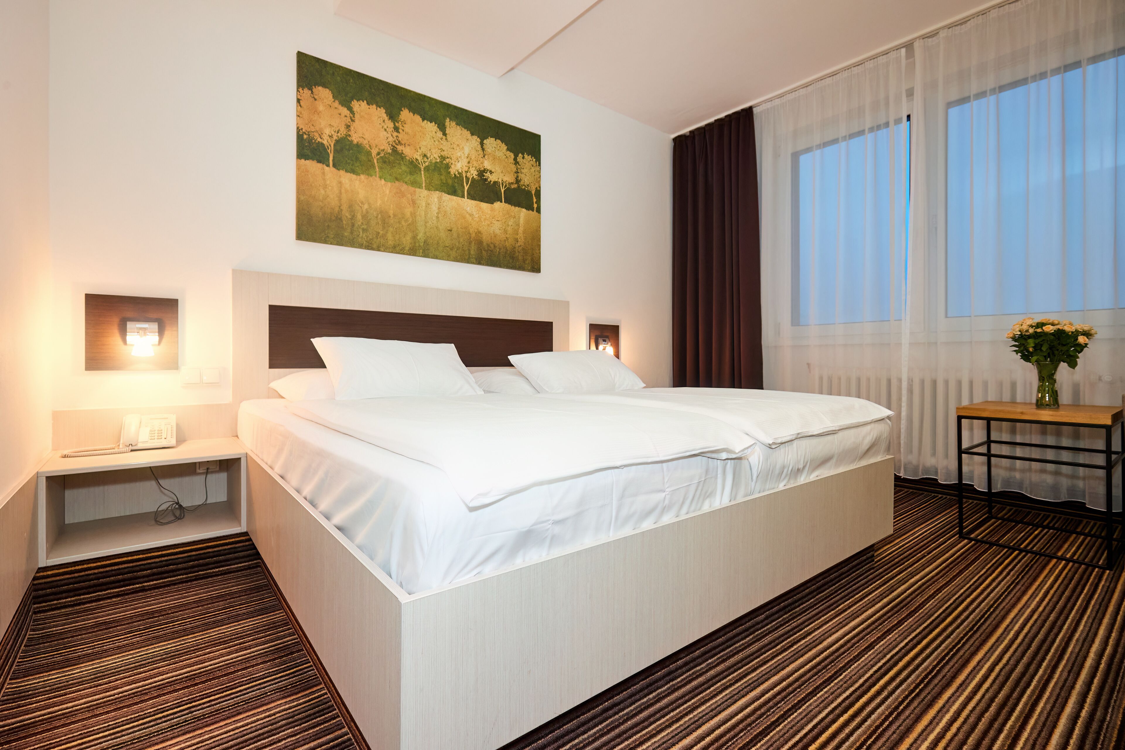 A hotel room with a large bed, bedside tables with lamps, a painting on the wall, a window with curtains, and a small table with a vase.