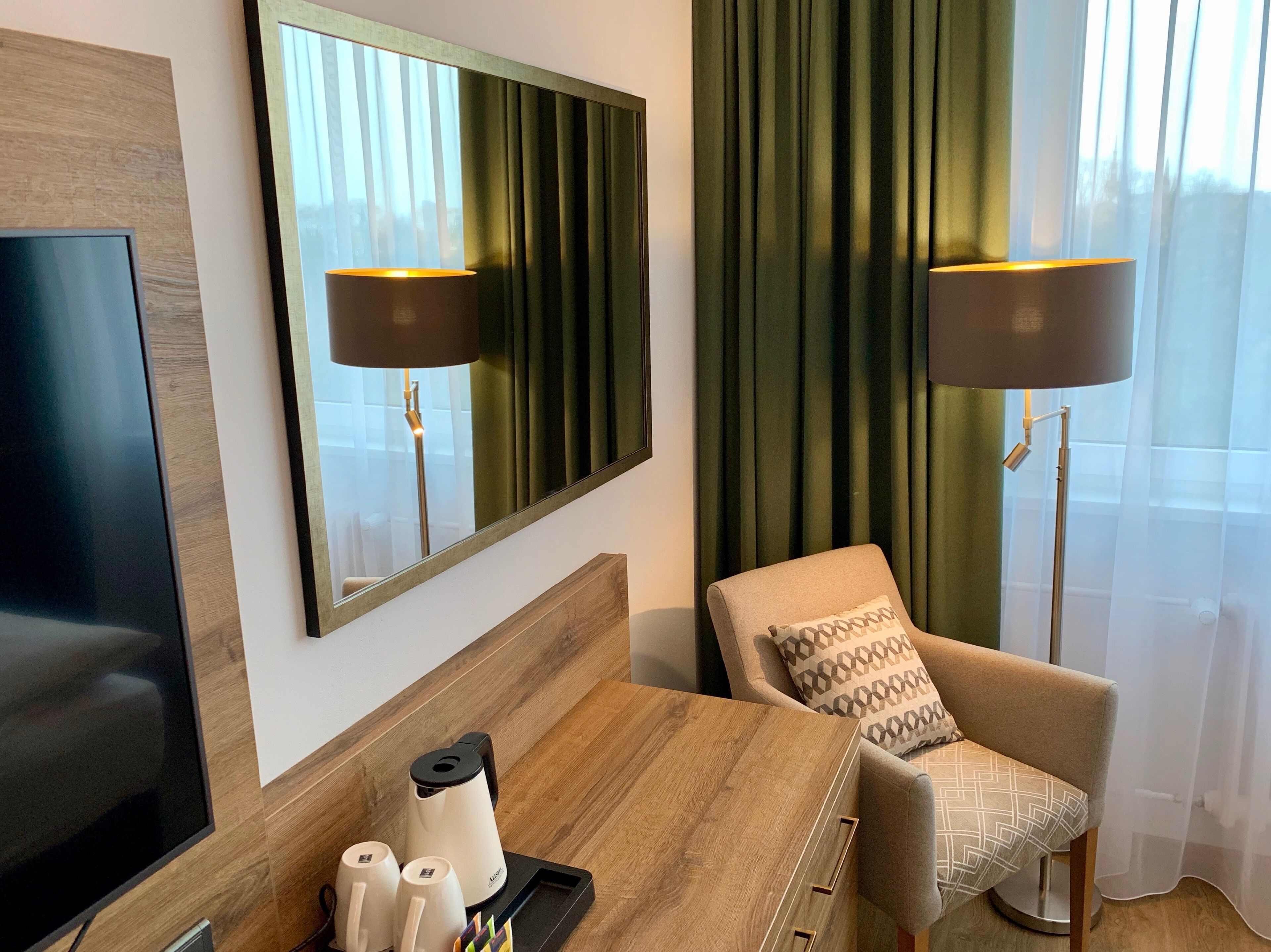 A hotel room with a wooden headboard, a flat-screen TV, a desk with a coffee maker, cups, and a tray, a patterned armchair, and two floor lamps.