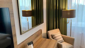 A hotel room with a wooden headboard, a flat-screen TV, a desk with a coffee maker, cups, and a tray, a patterned armchair, and two floor lamps.