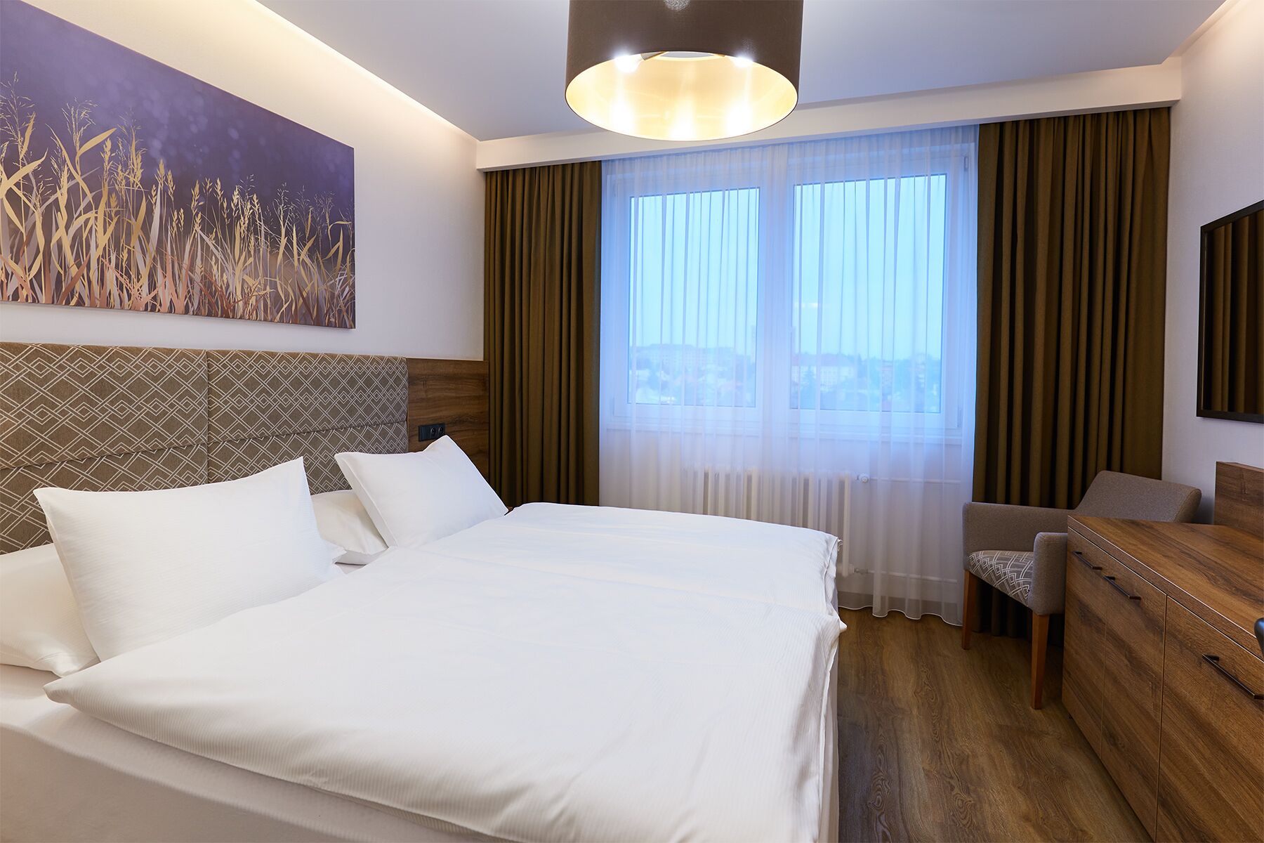 A hotel room with a large bed, a wooden headboard, a desk, and a chair. There is a painting on the wall and a window with curtains.