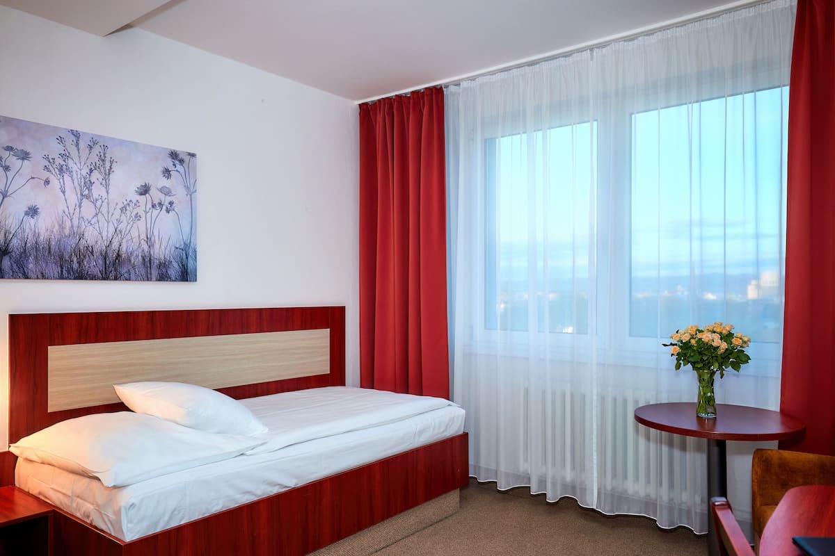 Een hotelkamer met een bed met houten hoofdeinde, witte beddengoed, een rood gordijn, een raam met uitzicht en een kleine tafel met een bloemenvaas.