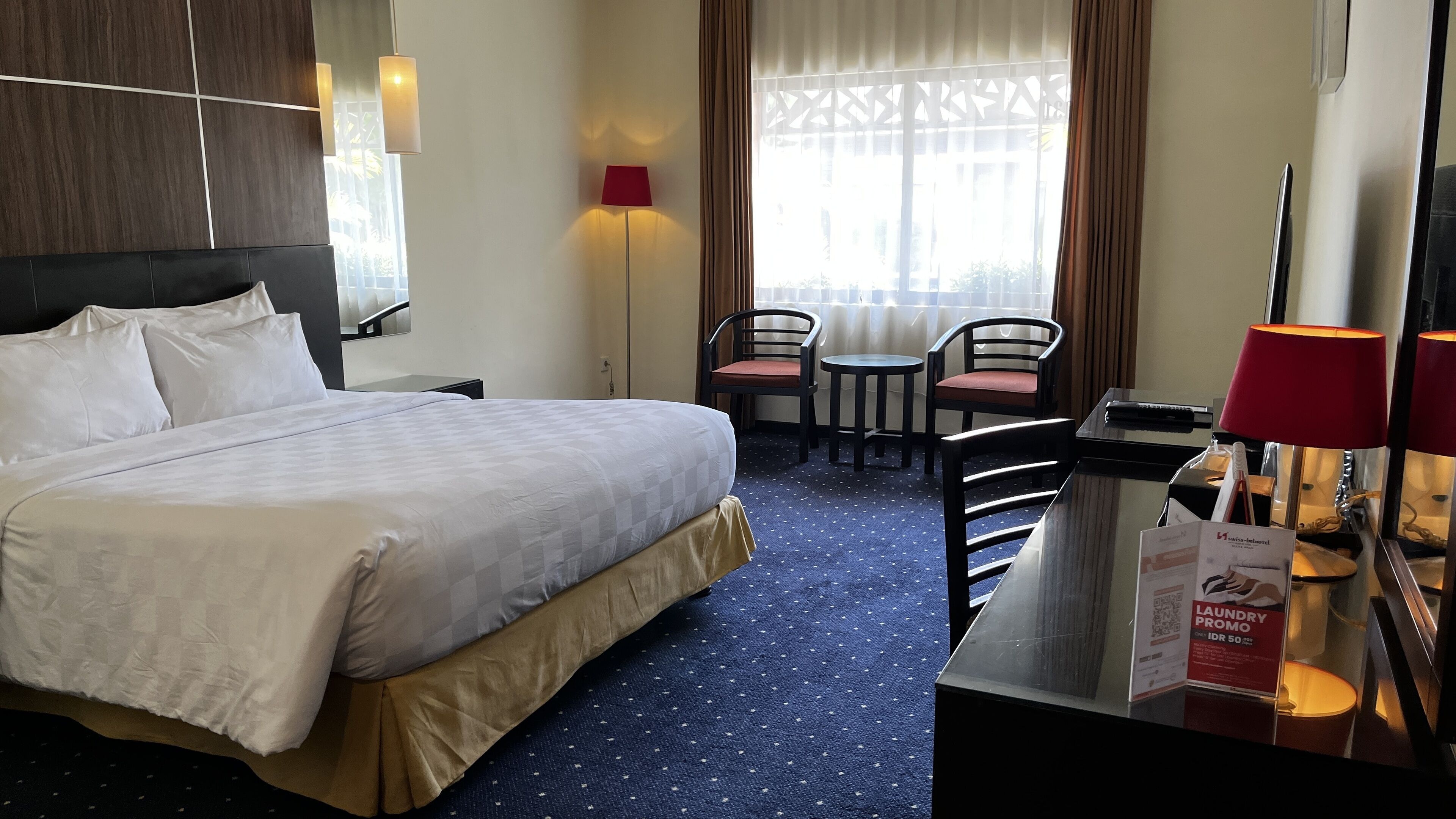 A hotel room with a bed, a desk, a chair, a lamp, and a window with curtains.