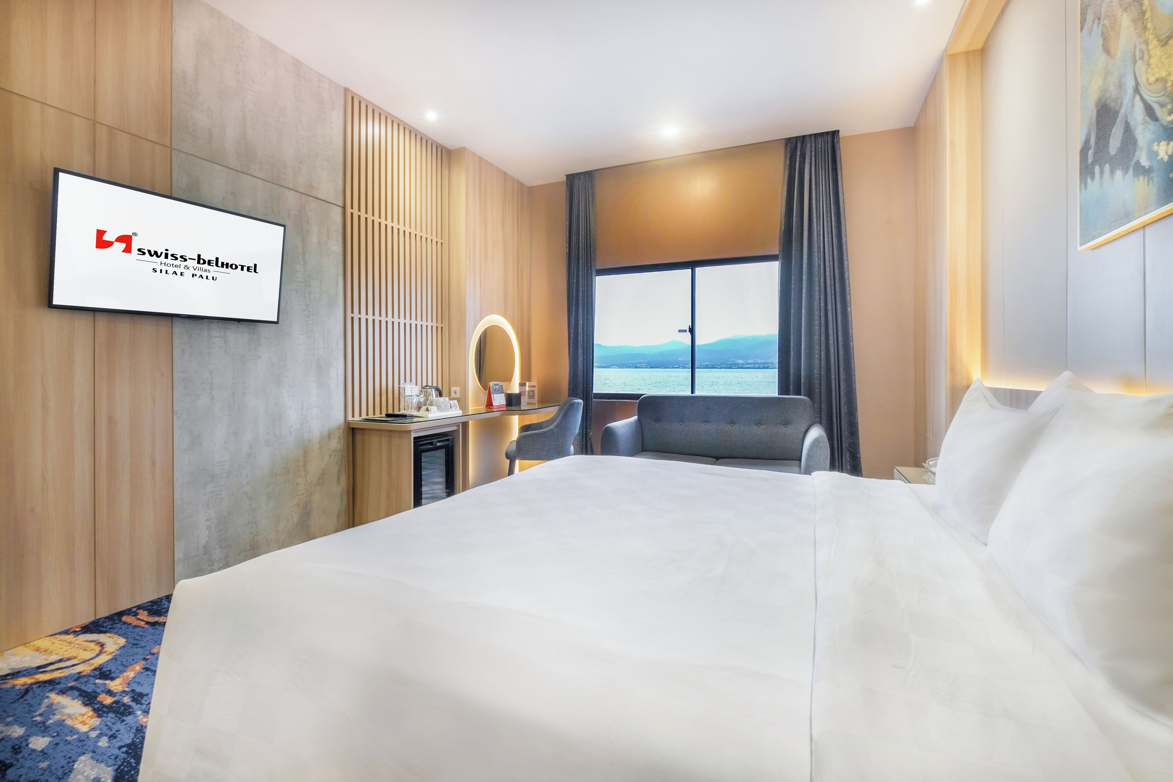 Habitación de hotel con una cama grande, un escritorio con silla, una mesita y un espejo, con vista al agua.