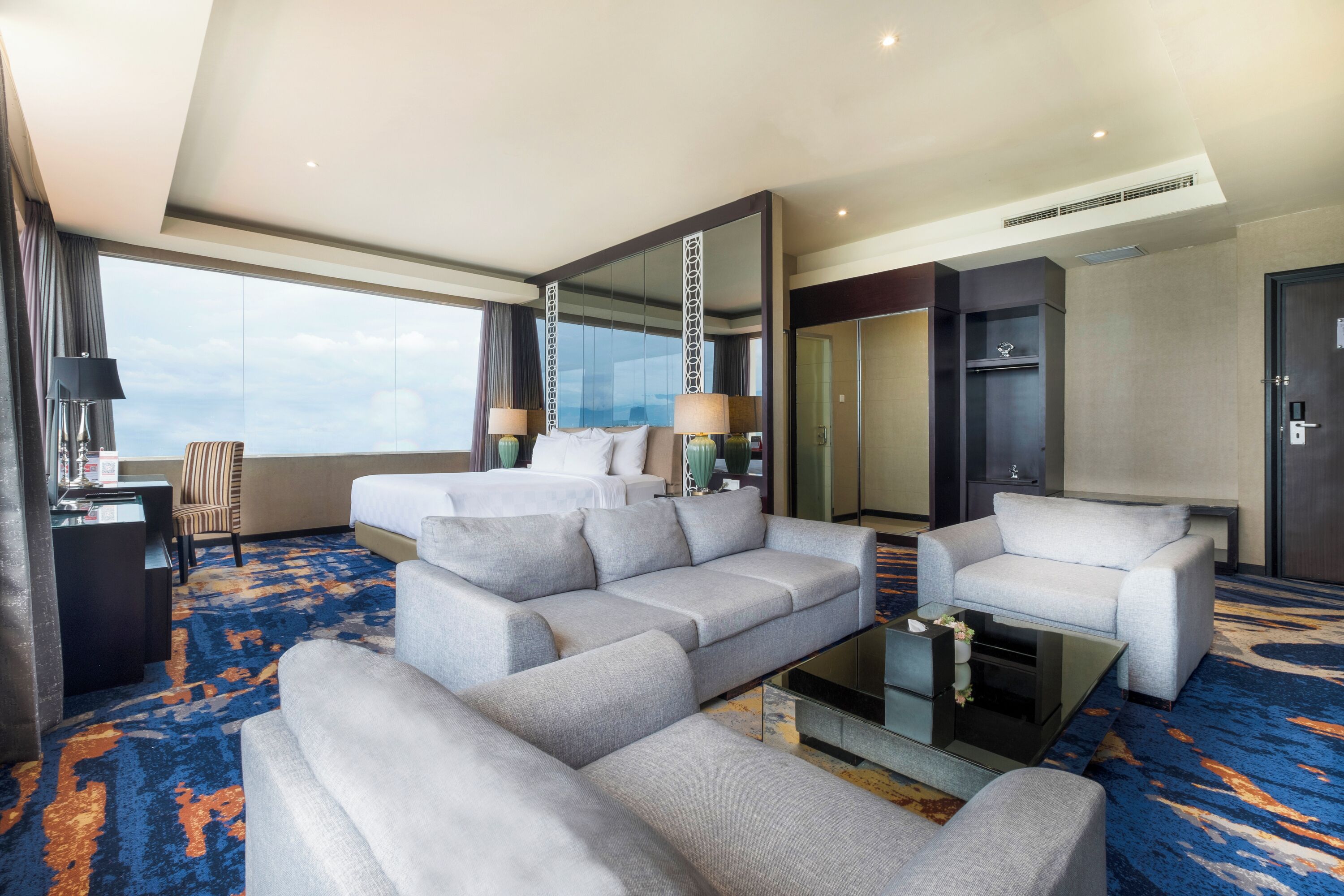 A modern hotel room with a large sectional sofa, a smaller sofa, a glass coffee table, and a desk with a lamp. There is a large mirror and a wardrobe. The room has a large window offering a view of the sky.