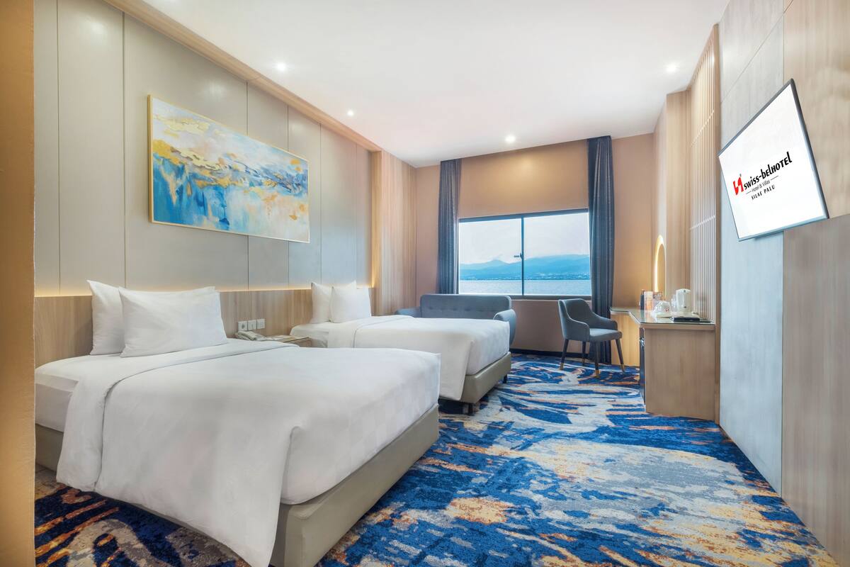 A hotel room with two beds, a desk, a chair, a large window, and a painting on the wall.