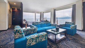 Een moderne woonkamer met blauwe banken en een salontafel, grote ramen met uitzicht op zee, en een kamerbreed tapijt met een patroon.