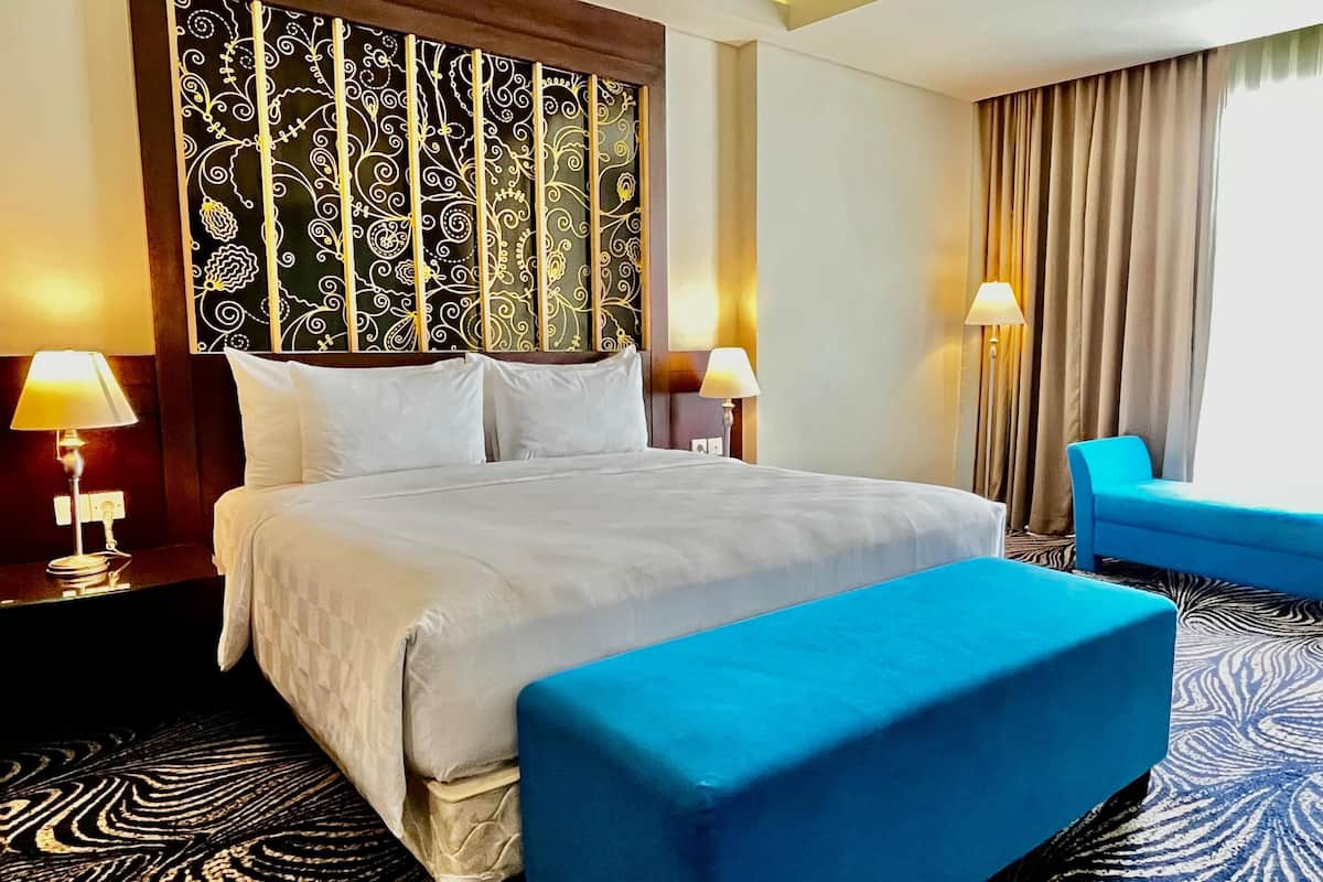 A hotel room with a large bed, two bedside tables with lamps, a blue armchair, a matching ottoman, a patterned carpet, and a decorative headboard.