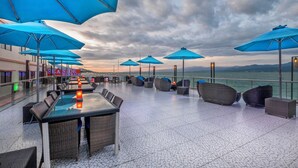 Een terras op het dak met blauwe parasols, rieten meubels en uitzicht op zee.