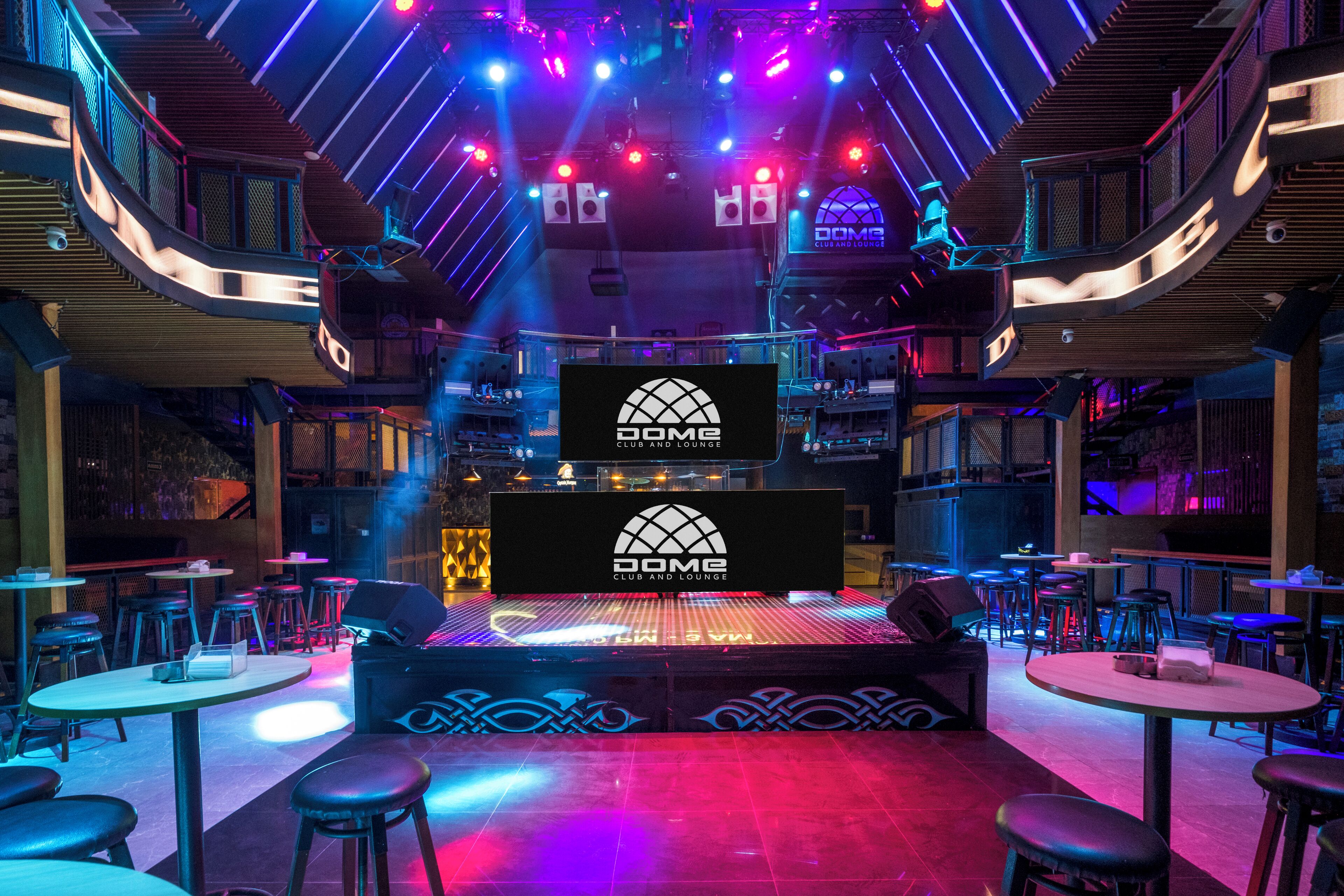 A modern lounge with a central stage, multiple seating areas, and a prominent logo on the stage.