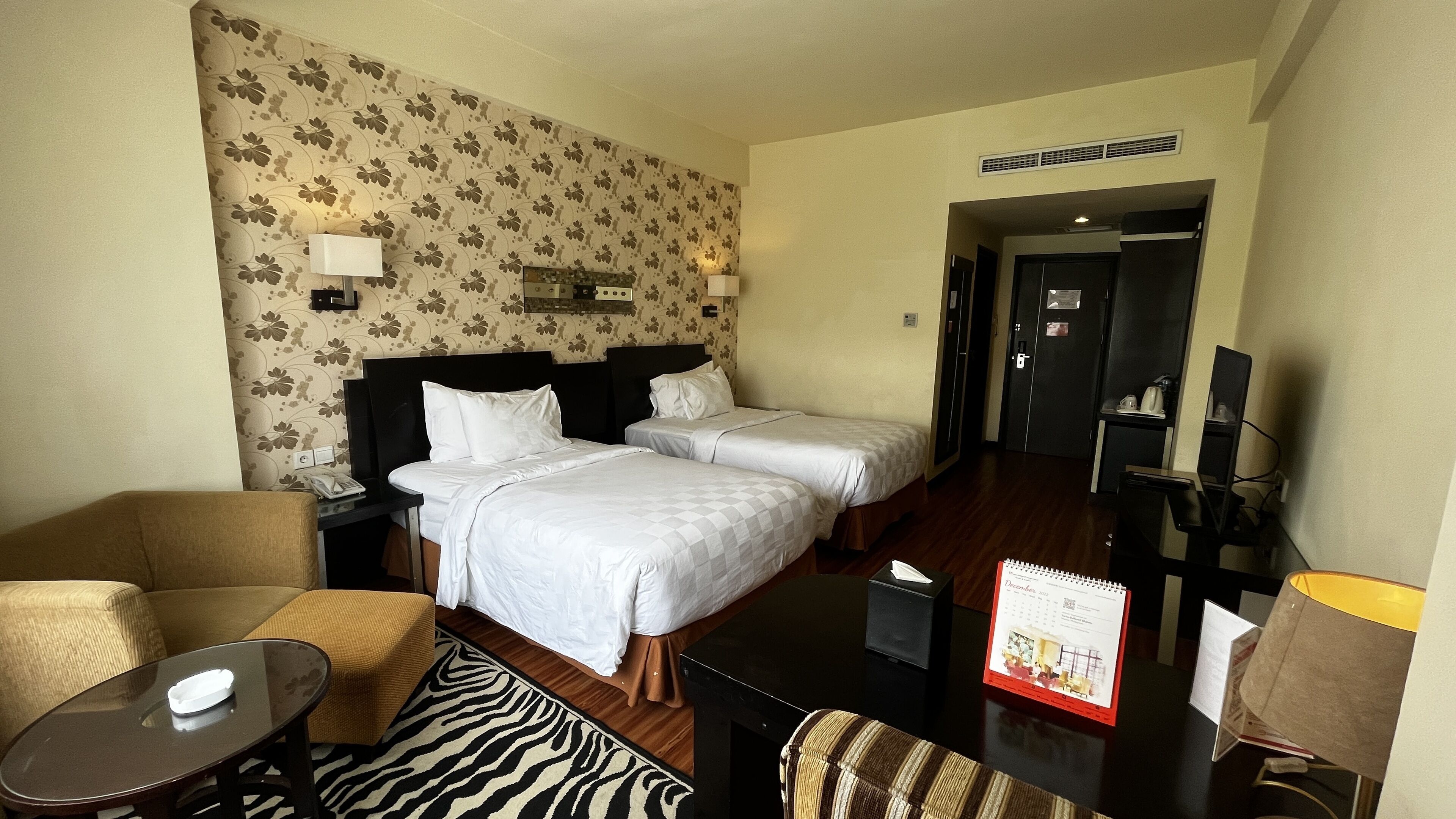 Quarto de hotel com duas camas, uma mesinha, uma cadeira, um tapete estampado com padrão de zebra e um calendário sobre a mesa.