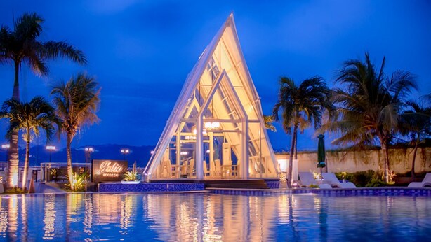 Een modern hotel met een glazen, driehoekige structuur, een zwembad en palmbomen.