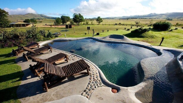 A swimming pool with wooden loungers and a pebble border, surrounded by a grassy landscape and distant hills. - Hawane Resort (Motshane)