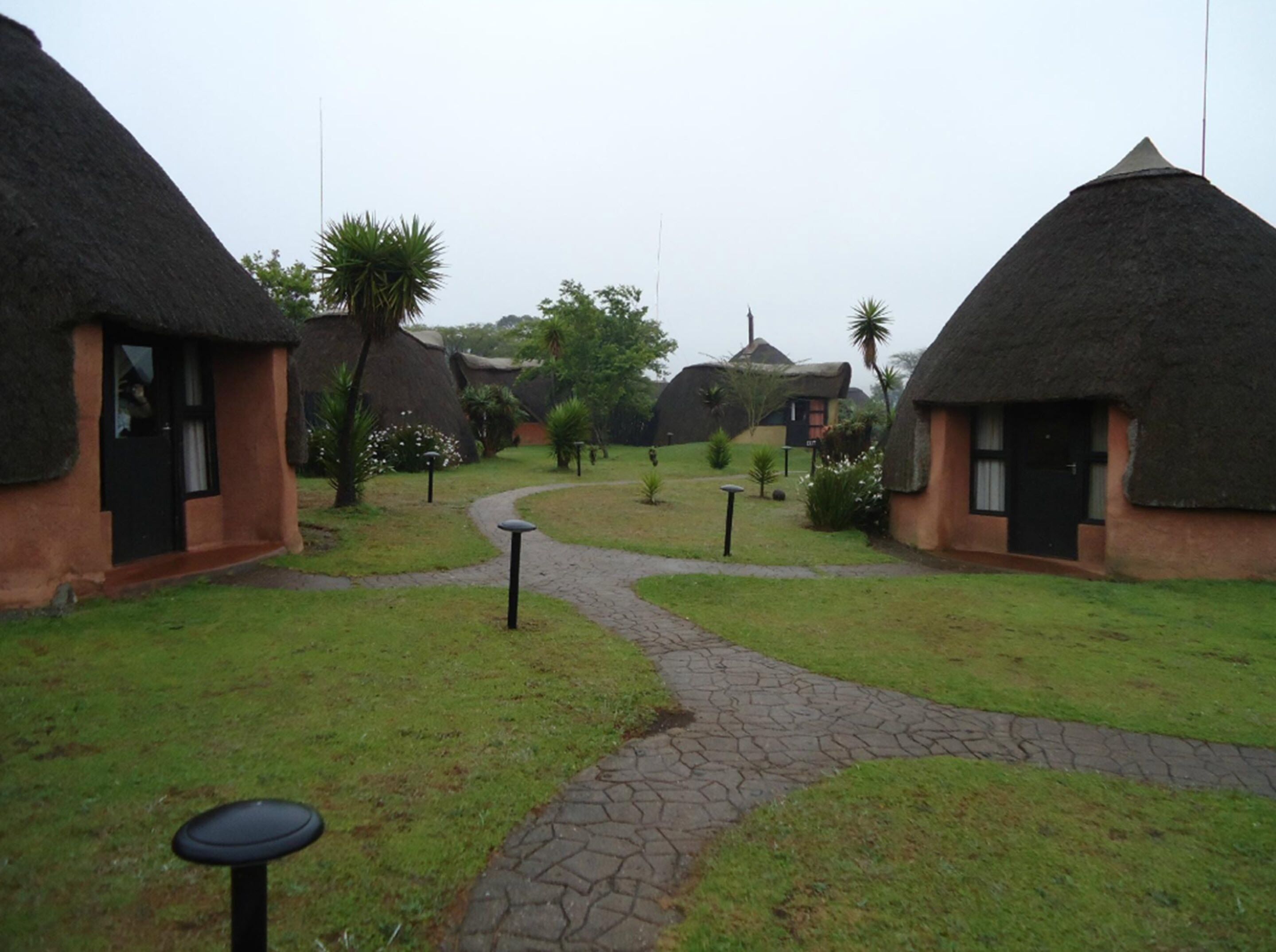 A pathway leading through a courtyard with thatched-roof huts and a small garden area.