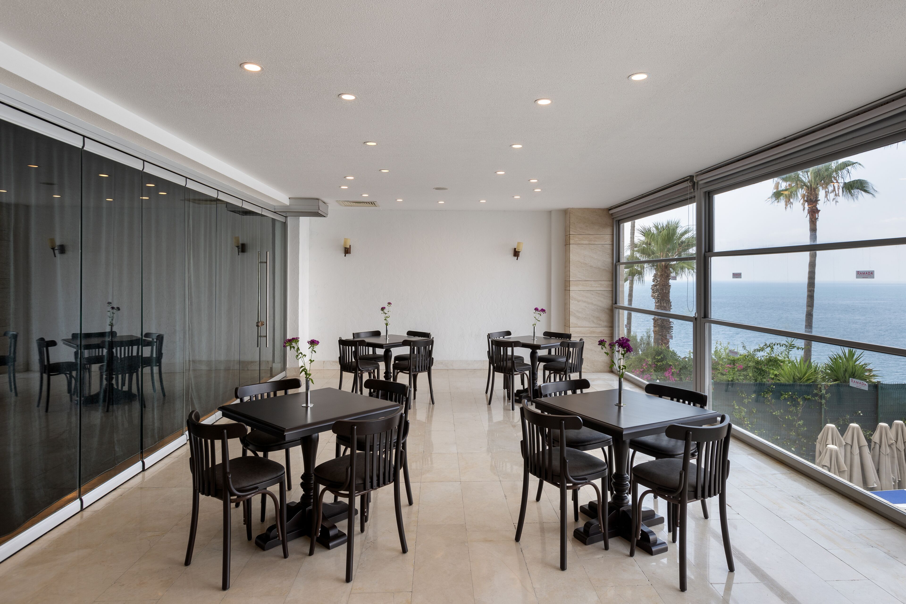 Un espace repas moderne avec des murs en verre, de nombreuses tables et chaises, offrant une vue panoramique sur l’océan.