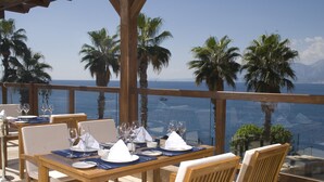 A dining area with tables set for two, overlooking a scenic ocean view with palm trees and mountains in the distance. - Ramada Plaza by Wyndham Antalya (Antalya)